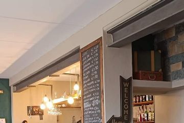 Bancone del bar con luci accese, piano in legno e scaffali con bottiglie esposte, lavagna e tavolino a forma di botte.