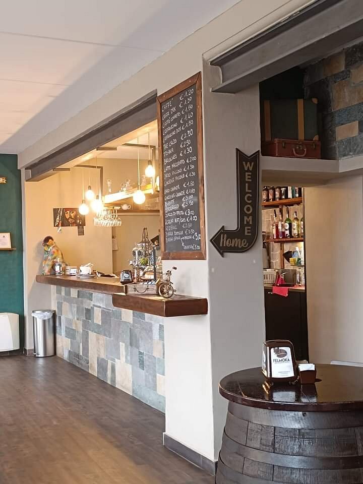 Bancone del bar con luci accese, piano in legno e scaffali con bottiglie esposte, lavagna e tavolino a forma di botte.